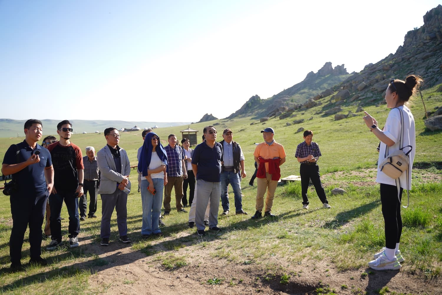 A group of people stands outdoors on a grassy landscape while a woman at right gestures and speaks to them under clear skies, engaging in discussion about Advancing Peace at the Mongolia Forum.