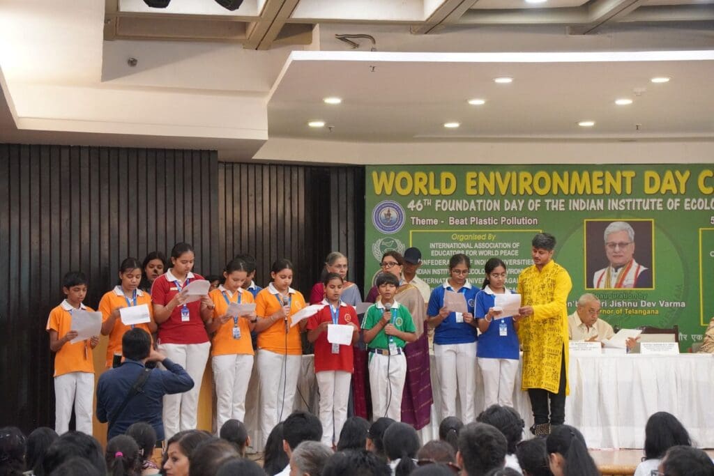 A group of students in colorful uniforms stand on stage reading from papers during the World Environment Day 2025 event, their global voices inspiring the audience seated before them to work together for a greener tomorrow.