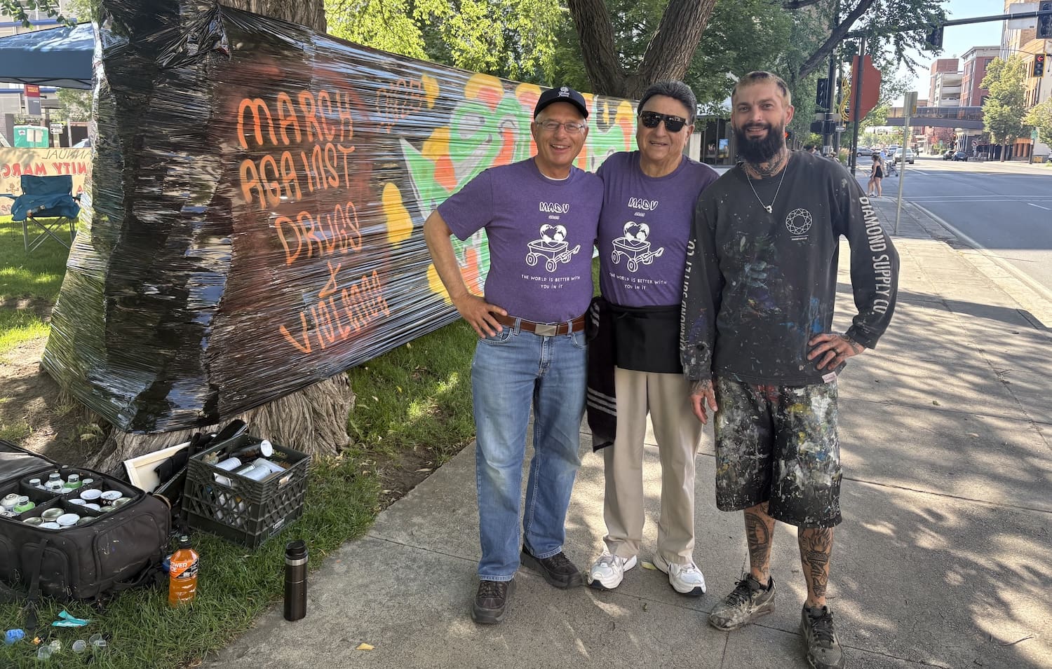 Three men stand on a sidewalk in front of a colorful mural and art supplies, showing support for grassroots peace—two wear matching purple shirts, while one is dressed in black paint-splattered clothing.
