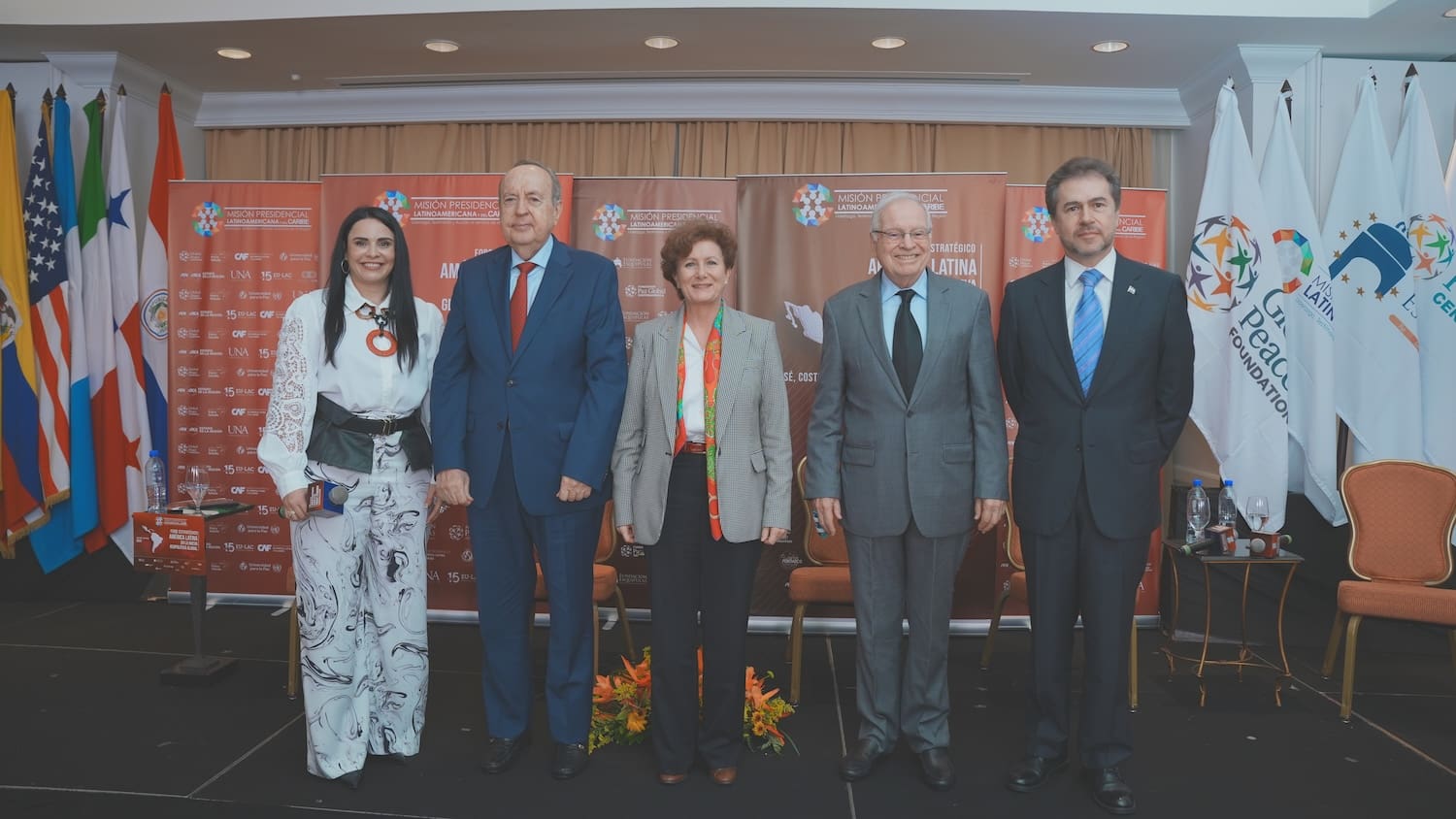 Five formally dressed individuals stand on a stage in front of international flags, with event branding that highlights Latin America's role in global geopolitics.