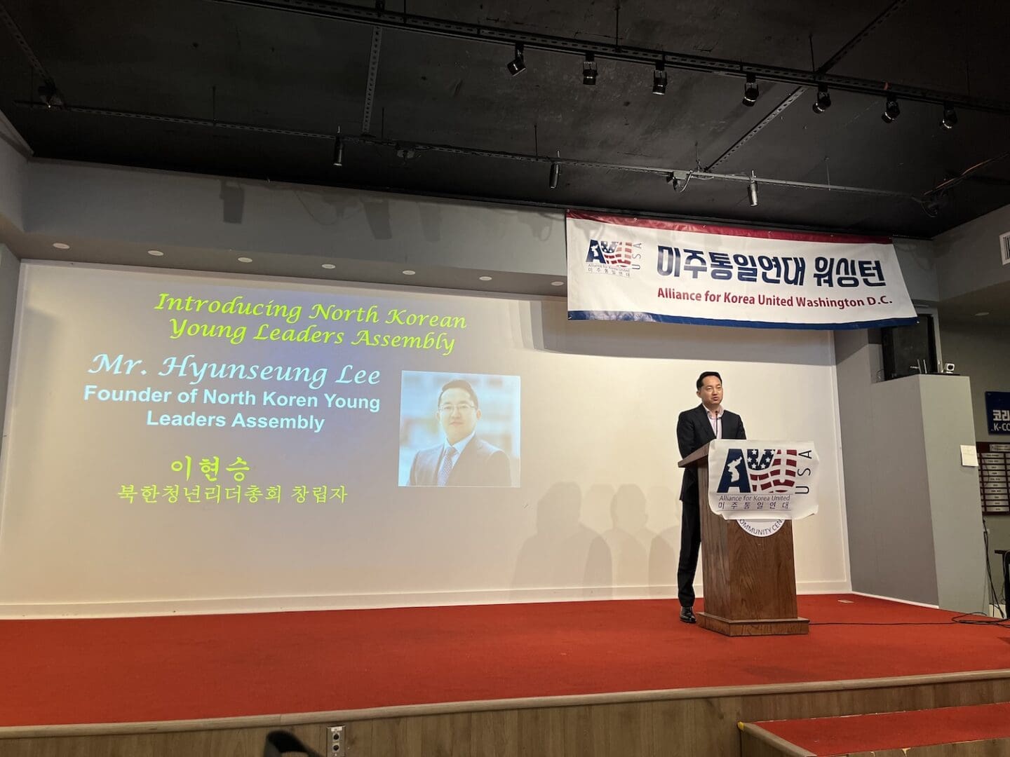 A man stands at a podium giving a presentation on the North Korean Young Leaders Assembly, with his photo and name, Mr. Hyunseung Lee, displayed prominently on the screen behind him.