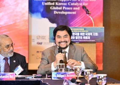 Two men in suits sit at a conference table with microphones; one smiles and speaks into a mic, while a colorful One Korea 2025 International Forum Day 2 event banner is visible in the background.
