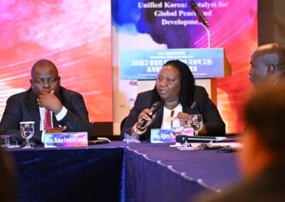 Three people participate in a panel discussion at the International Forum on One Korea, with one woman speaking into a microphone and a banner displayed in the background.