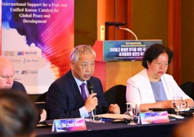 On Day 2 of the International Forum, three panelists sit at a conference table with microphones, speaking and taking notes. A banner behind them highlights support for a free and unified Korea under the One Korea 2025 initiative.