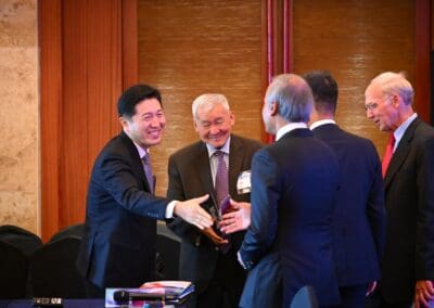 Five men in business attire stand indoors, smiling and greeting each other at the International Forum on One Korea; one man extends his hand for a handshake across a table with documents.