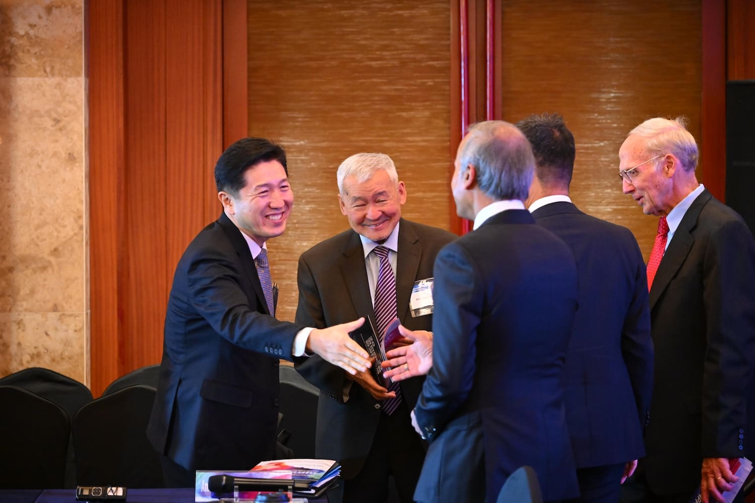 Five men in business attire stand indoors, smiling and greeting each other at the International Forum on One Korea; one man extends his hand for a handshake across a table with documents.
