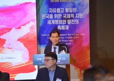 A man in a suit speaks at a podium during the International Forum on One Korea, with a colorful backdrop displaying Korean and English text about international support for a free and unified Korea.