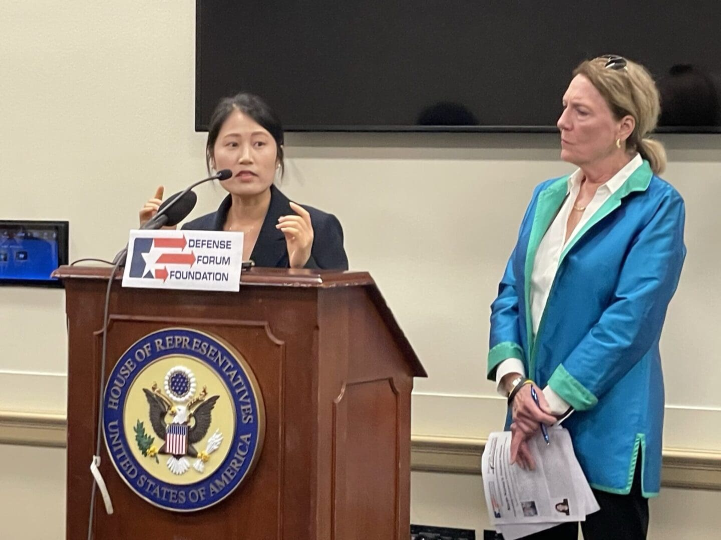 A woman speaks at a podium with the House of Representatives seal, while another woman stands nearby holding papers, during a Defense Forum Foundation event for the North Korean Young Leaders Assembly.