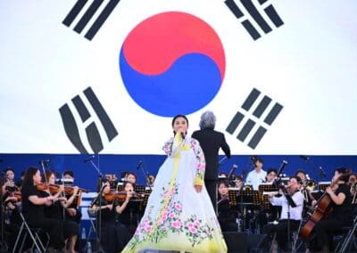 A woman in a traditional Korean dress sings in front of an orchestra, with a large South Korean flag displayed in the background, celebrating the spirit of the Korean Dream at the 2025 Hangang Festa.