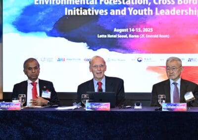 Three men in suits sit at a conference panel table with microphones and nameplates, in front of a screen displaying event details about environmental forestation, youth leadership, and the International Forum on One Korea.
