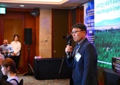 A man speaks into a microphone at a conference. Behind him, a large screen displays “Keep Mongolia Green Project” alongside a forested landscape, highlighting the initiative during the International Forum on One Korea.