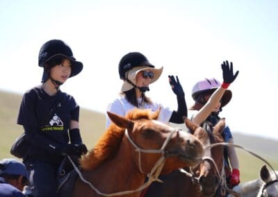 Three people wearing helmets ride horses outdoors; two wave their hands while the third looks ahead, evoking a spirit of Northeast Asian Peaceful Development against a clear and sunny background.