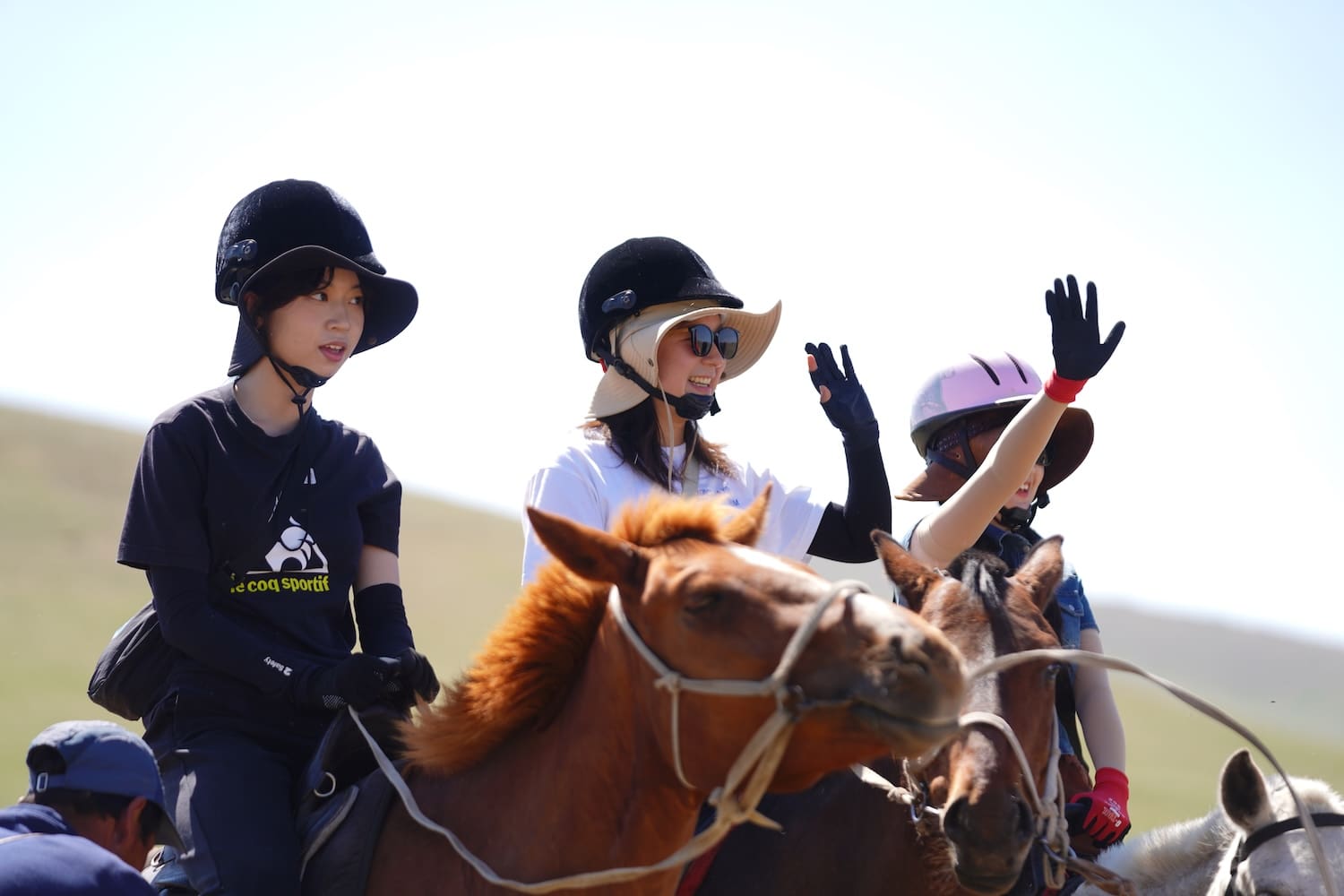 Three people wearing helmets ride horses outdoors; two wave their hands while the third looks ahead, evoking a spirit of Northeast Asian Peaceful Development against a clear and sunny background.