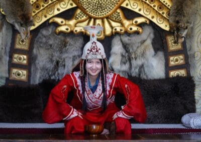 A person in traditional red attire and an ornate headdress sits indoors in front of a decorated wall with fur and golden patterns, smiling at the camera—evoking the spirit of Mongolia Forum and Northeast Asian development.