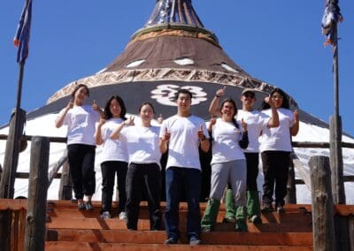 Seven people in white shirts stand on wooden steps in front of a large round structure, all giving a thumbs-up gesture under a clear blue sky, symbolizing support for Korean Unification and Northeast Asian Peaceful Development.