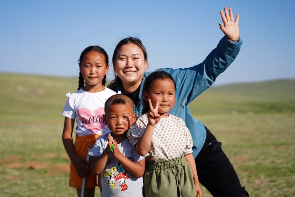 Four children and a young woman pose outdoors on a sunny day, with the woman waving and the group smiling at the camera against a grassy landscape—moments like these often shared at events such as the Mongolia Forum.