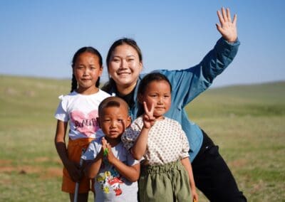 Four children and a young woman pose outdoors on a sunny day, with the woman waving and the group smiling at the camera against a grassy landscape—moments like these often shared at events such as the Mongolia Forum.