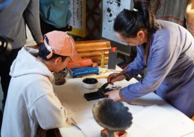 A woman demonstrates calligraphy techniques to a seated man at a table with ink, brushes, and paper, as others observe—capturing the spirit of Northeast Asian Peaceful Development at the 2025 Mongolia Forum.