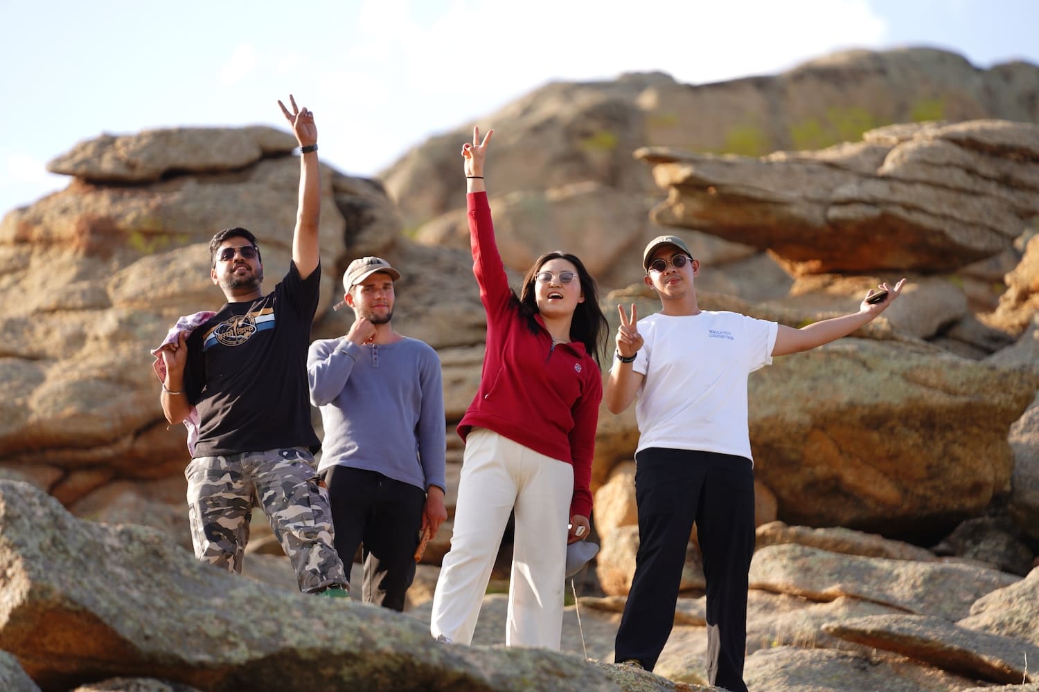 Four people stand on rocky terrain, facing forward and making peace signs; the backdrop of large rocks and blue sky hints at a shared hope for Northeast Asian development or Korean unification.