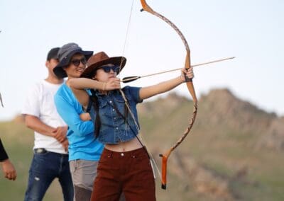A person aims a traditional bow and arrow outdoors, with three others standing nearby on a grassy field with hills in the background—an image evoking themes of Northeast Asian Peaceful Development.