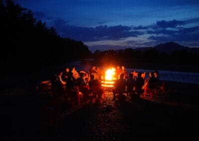 A group of people sit on red chairs around a large campfire at dusk near a river, discussing Northeast Asian development, with trees and mountains silhouetted in the background.