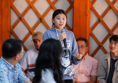 A young woman holding a microphone speaks to a seated group of people indoors, with wooden paneling on the wall behind them at a Mongolia Forum discussing Northeast Asian Peaceful Development.