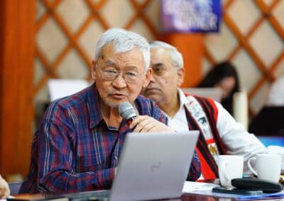 An older man speaks into a microphone at a table with a laptop and mugs, while another man sits beside him, during a discussion on Northeast Asian Peaceful Development at the 2025 Mongolia Forum.
