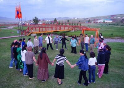 A group of people stands in a large circle holding hands on a grassy field, representing unity and hope for Korean Unification, with buildings and flags visible in the background.