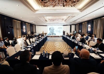 A conference room with attendees seated around a U-shaped table, watching a presentation projected onto a screen showing a portrait of a man at the Mongolia forum on Northeast Asian peaceful development.