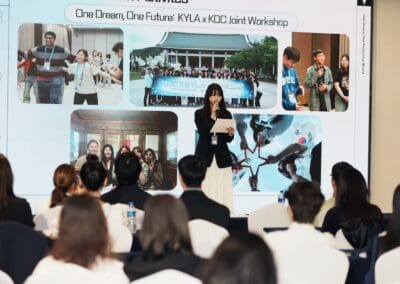 A woman stands at a podium presenting to an audience, with a slideshow behind her displaying group photos from a joint workshop event focused on Korean Unification and Northeast Asian Peaceful Development.