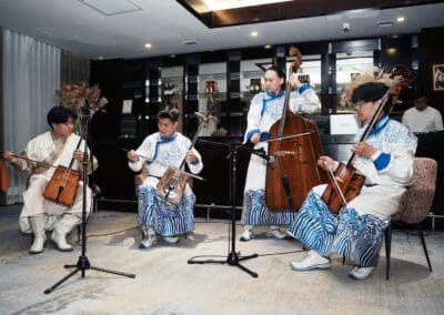 Four musicians in traditional attire perform string instruments on stage at a modern indoor venue, embodying the spirit of Northeast Asian Peaceful Development. A person stands behind a counter in the background.