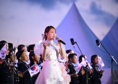 A woman in a white dress sings into a microphone on stage at Hangang Festa 2025, accompanied by children holding South Korean flags, expressing the spirit of the Korean Dream with white tents in the background.