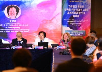 Three panelists sit at a table with microphones during the 2025 One Korea International Forum, with a large banner and a projected image of a woman in the background. Audience members are visible in the foreground.