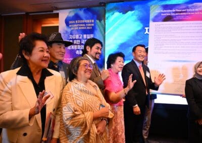 A group of people in formal attire smile and wave while standing in front of colorful posters at the One Korea 2025 International Forum event.