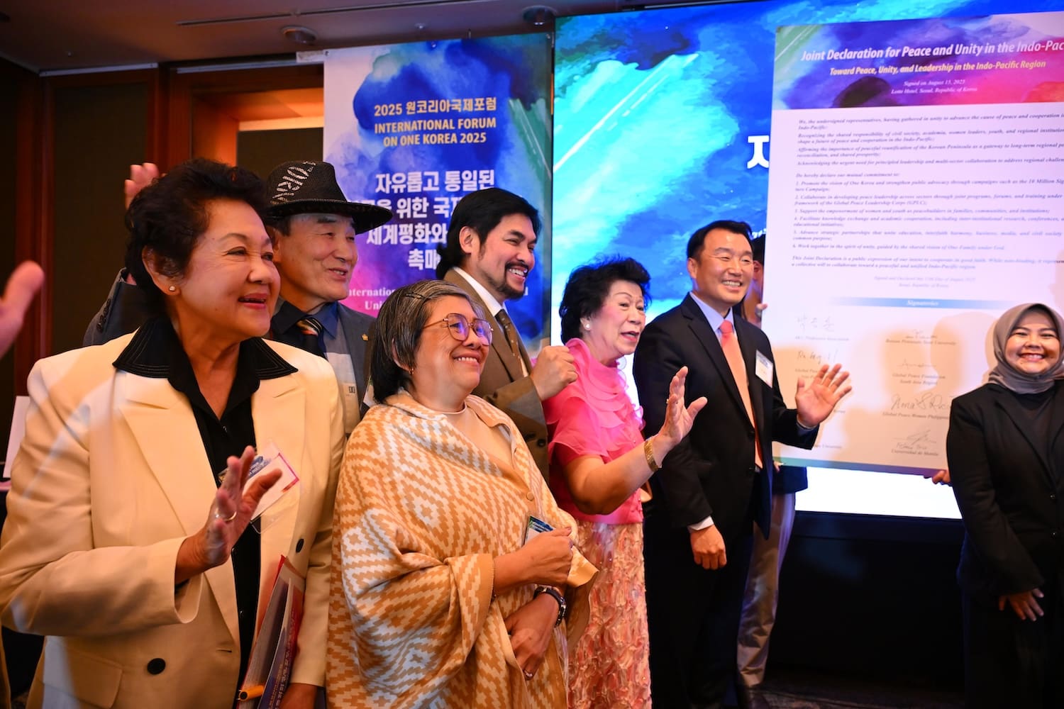 A group of people in formal attire smile and wave while standing in front of colorful posters at the One Korea 2025 International Forum event.