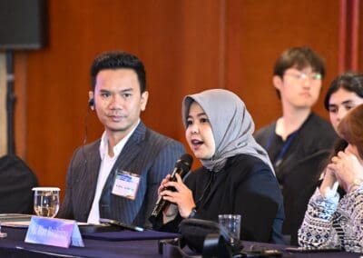 At the International Forum 2025, a woman in a hijab speaks into a microphone at a conference table, while others listen attentively—highlighting the spirit of unity around the One Korea initiative.