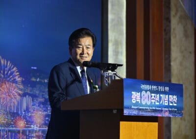 A man in a suit speaks at a podium during an event commemorating the National Liberation Day of Korea, set against a cityscape and fireworks backdrop at the International Forum on One Korea.