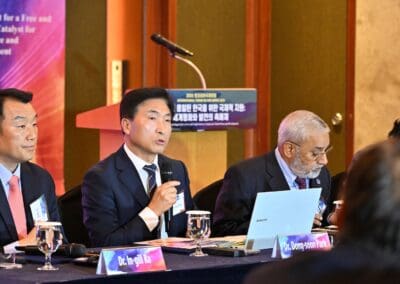 Three men in suits sit at a conference table with microphones, laptops, and nameplates. The man in the center speaks into a microphone. Banners for the 2025 International Forum on One Korea are visible in the background.