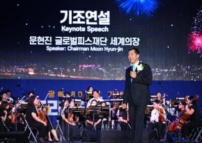 A man stands on stage holding a microphone in front of an orchestra, delivering a keynote speech introduction as fireworks and the "Korean Dream" light up the screen behind him at the 2025 Hangang Festa.