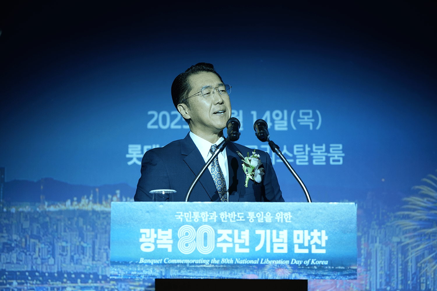 A man in a suit speaks at a podium during a banquet commemorating the 80th National Liberation Day of Korea, with cityscape and event details in the background.