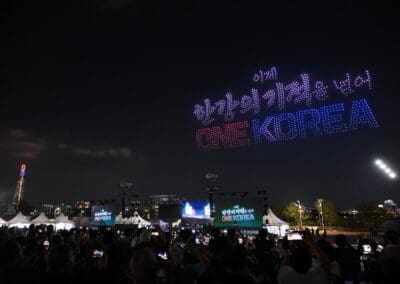 A nighttime crowd at Hangang Festa 2025 watches a drone light show displaying Korean text and the words "ONE KOREA" in red and blue, inspiring all with the vision of a Korean Dream above the festive outdoor event.