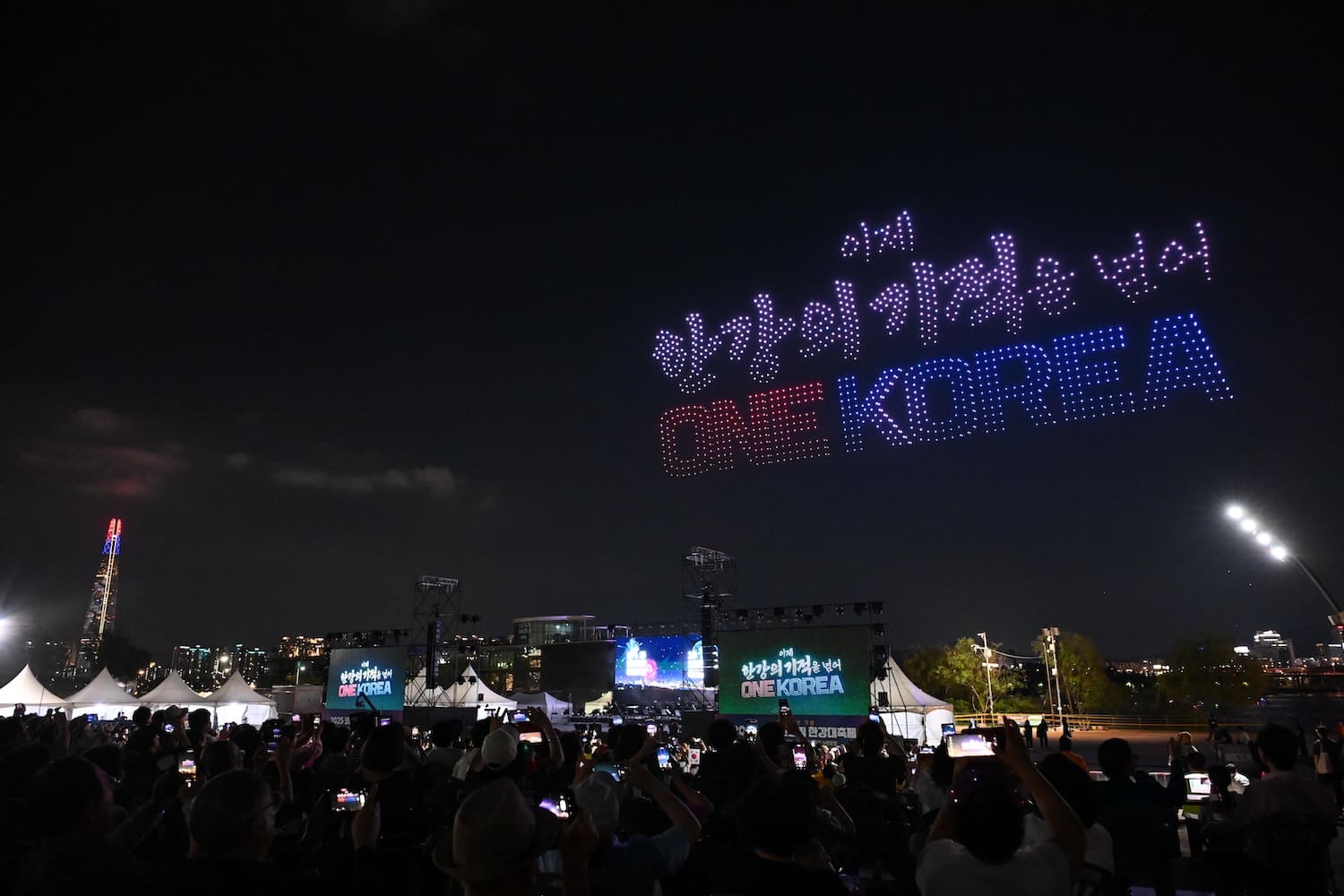 A nighttime crowd at Hangang Festa 2025 watches a drone light show displaying Korean text and the words