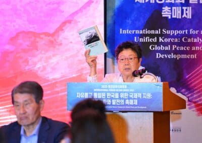 An older woman speaks at a podium, holding up a book, with a banner behind her displaying text about global peace and support for the International Forum on One Korea and Korean unification.