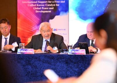 Three men in suits sit at a conference table, one speaking into a microphone, with banners and nameplates visible in the background at the 2025 International Forum on One Korea.