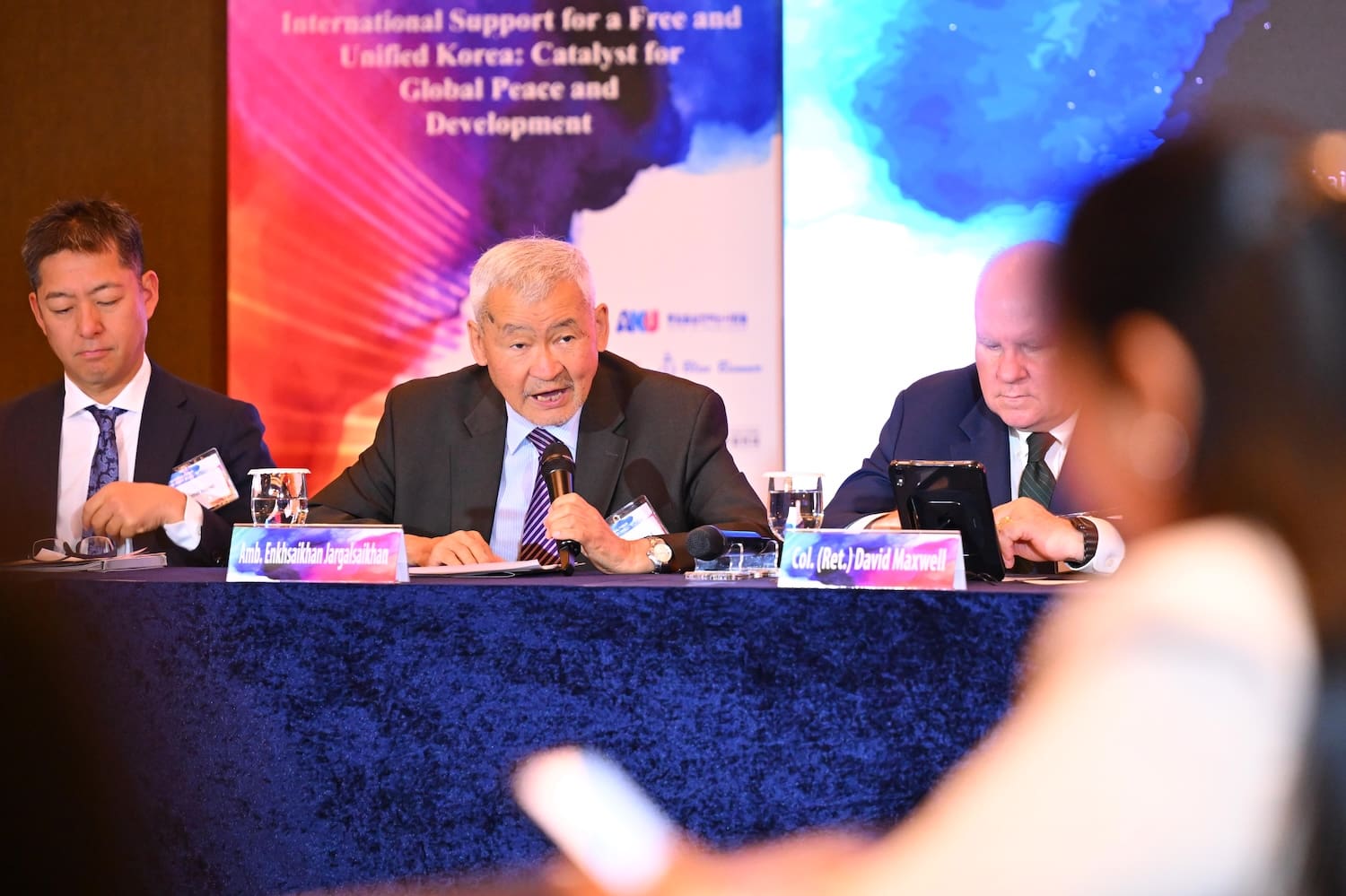 Three men in suits sit at a conference table, one speaking into a microphone, with banners and nameplates visible in the background at the 2025 International Forum on One Korea.