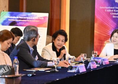 A woman speaks into a microphone during an International Forum panel discussion, seated at a table with nameplates, water glasses, and colorful One Korea 2025 posters in the background.