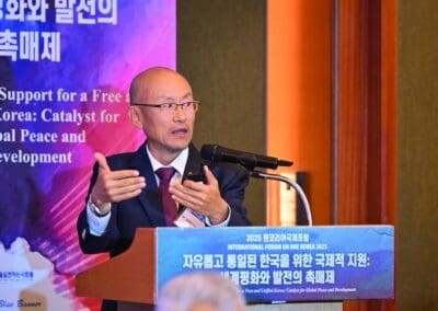 A man in a suit and glasses speaks at a podium during the 2025 International Forum on One Korea, with banners in Korean and English for the International Forum on One Korea displayed behind him.