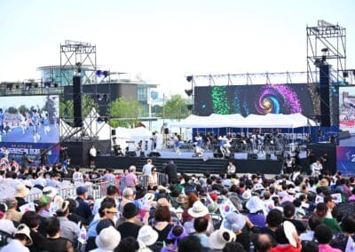 A large outdoor audience watches a stage with musicians and a colorful screen at the 2025 Hangang Festa, surrounded by lighting rigs and tents, during a public event on a sunny day.