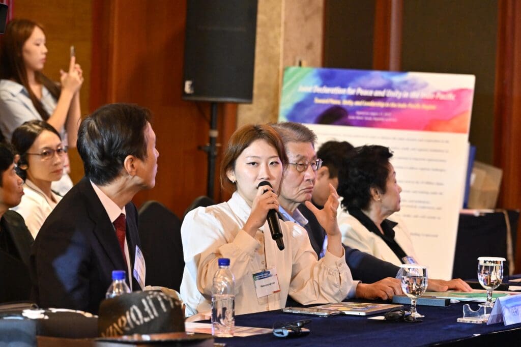 A woman speaks into a microphone at a conference table with other attendees during Day 2 of the International Forum, while an audience listens in the background.