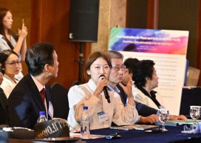 A woman speaks into a microphone at a conference table with other attendees during Day 2 of the International Forum, while an audience listens in the background.
